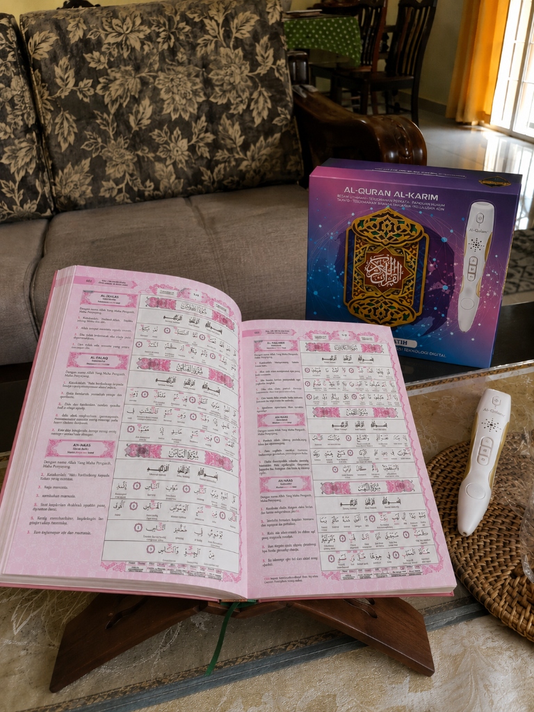 Gambar snap fon pelanggan: Quran A4 terbuka atas rehal kayu dengan kotak ungu & pen di ruang tamu